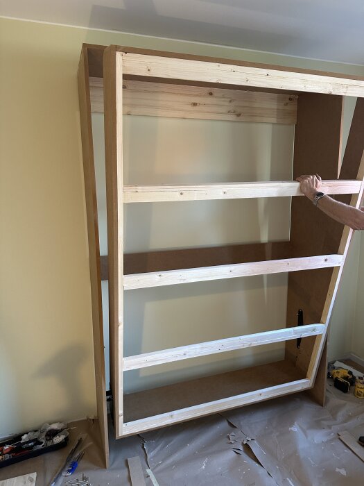 Träram till sängskåp under konstruktion, sedd inomhus med verktyg och material omkring, ljusgula väggar i bakgrunden.