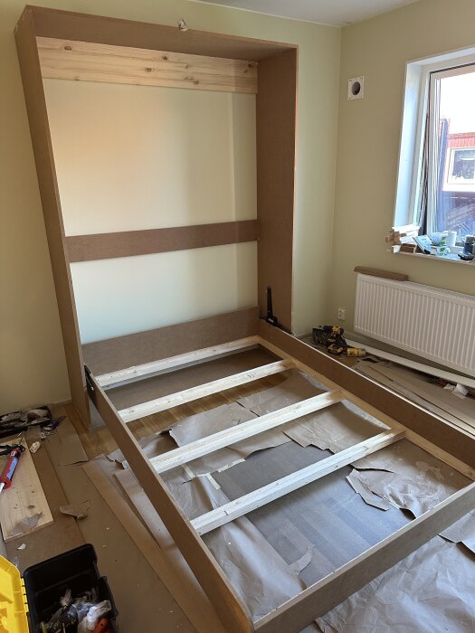 Hemmagjord sängram i trä under byggnation i ett DIY-projekt, med ställning för en Murphy Bed, verktyg och material runtom.