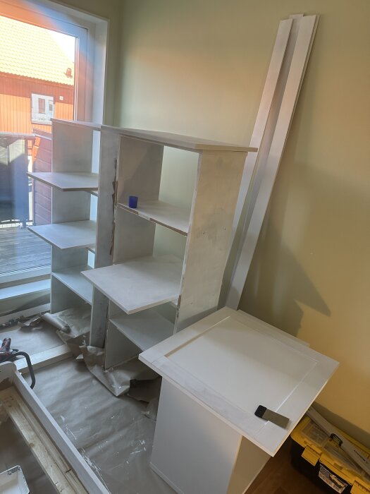 Vitt sängskåp under konstruktion i rum med ljusa väggar. Hyllor och skivor ligger mot väggen, och byggmaterial syns på golvet.