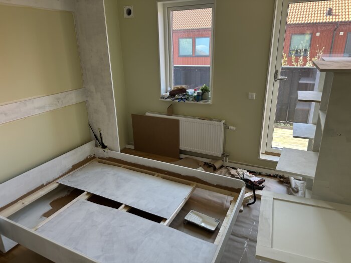 Murphy Bed under konstruktion, sängen är nedfälld i ett rum med gräddvita väggar och stora fönster. Material och verktyg syns på golvet.
