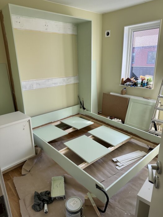 Pågående byggprojekt av ett sängskåp med synlig sängram i nedfällt läge, omgiven av verktyg och material i ett halvfärdigt rum.