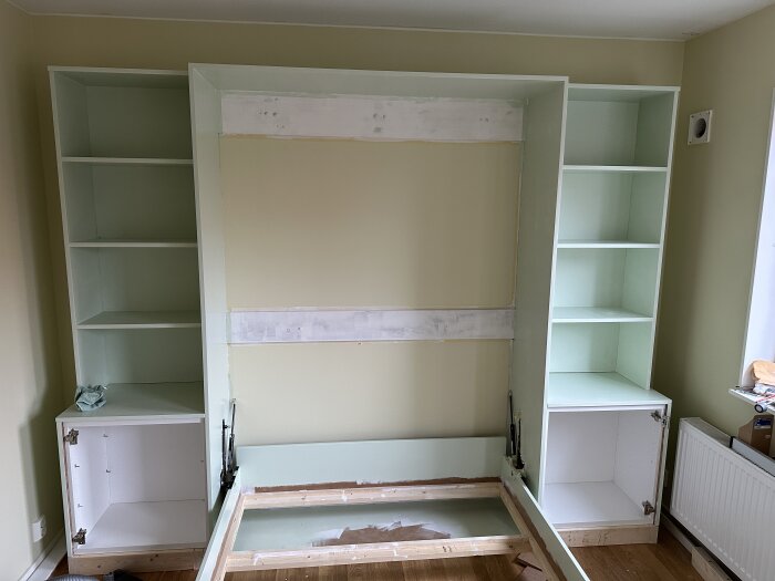 Nedfällt sängskåp byggt med MDF och hyllor, flankerat av skåp på vardera sida. Väggarna är nyligen målade. Inga socklar eller knoppar är monterade.
