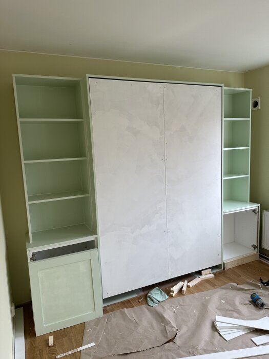 Inbyggt sängskåp med öppna hyllor på sidorna i ett rum under renovering, golvet täckt med papper och verktyg.