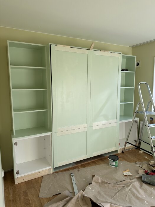 Grönt sängskåp under uppbyggnad, med monterade hyllor bredvid, stege och verktyg bredvid. Täckt golv för skydd mot färgstänk.