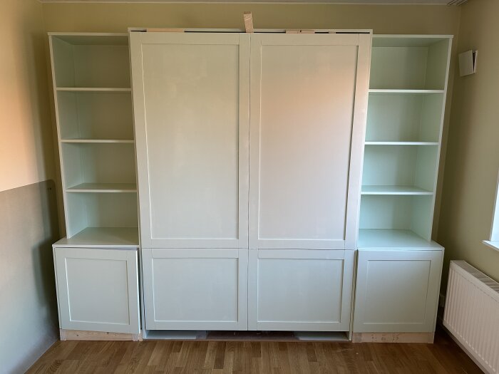 Inbyggt sängskåp med hyllor på både sidor, stående mot en vägg i ett rum. Skåpet har dubbeldörrar och omålade socklar.