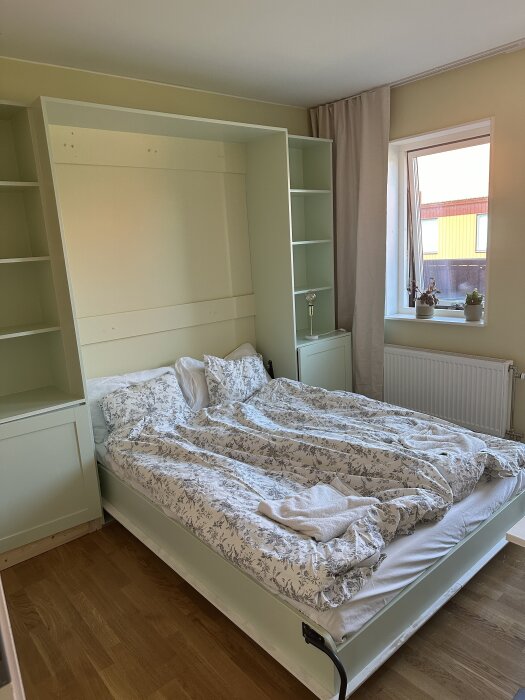 En Murphy säng med blommat bäddset är nedfälld i ett rum med grönmålade skåp och hyllor på sidorna, bredvid ett fönster med krukväxter.