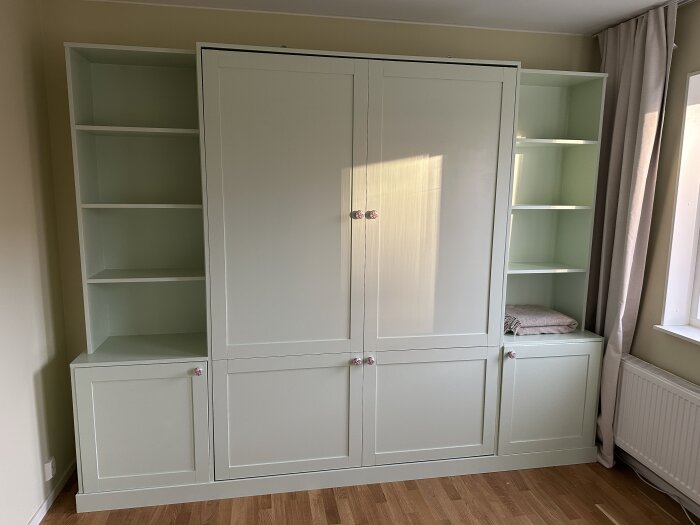 Grönt sängskåp med omgivande hyllor och skåp, står i ett rum med träfärgat golv och ljusa väggar.