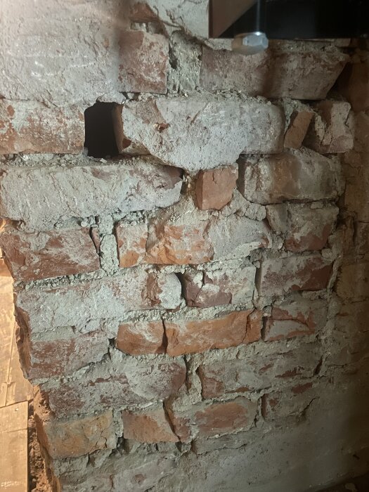 Skadad murstock med exponerade tegelstenar och ett större hål, delar av cement synliga.