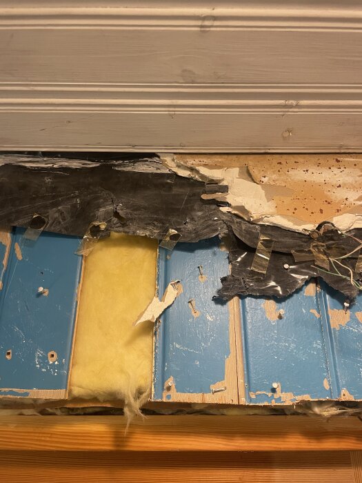 Svart plastfilm och gul isolering bakom blå panel; gipsrester syns ovanför, trolig tidigare renovering, pågående husprojekt.