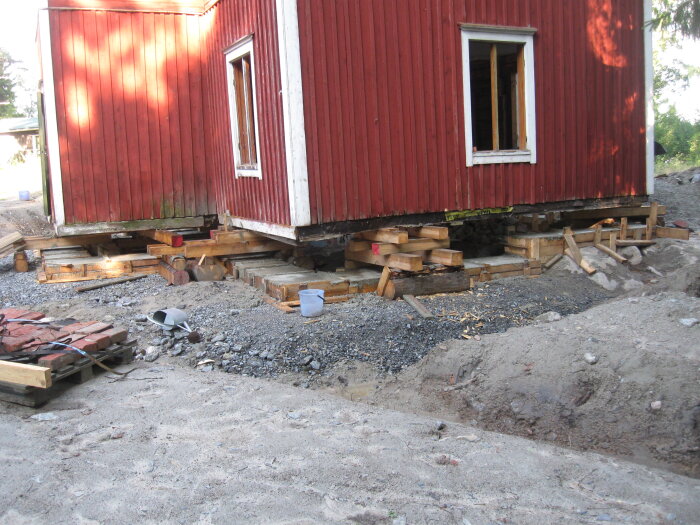 Rött hus på tillfälliga trästöddar i ett renoveringsprojekt, omringar av sand och grus. Huset är uppstöttat för grundarbete.