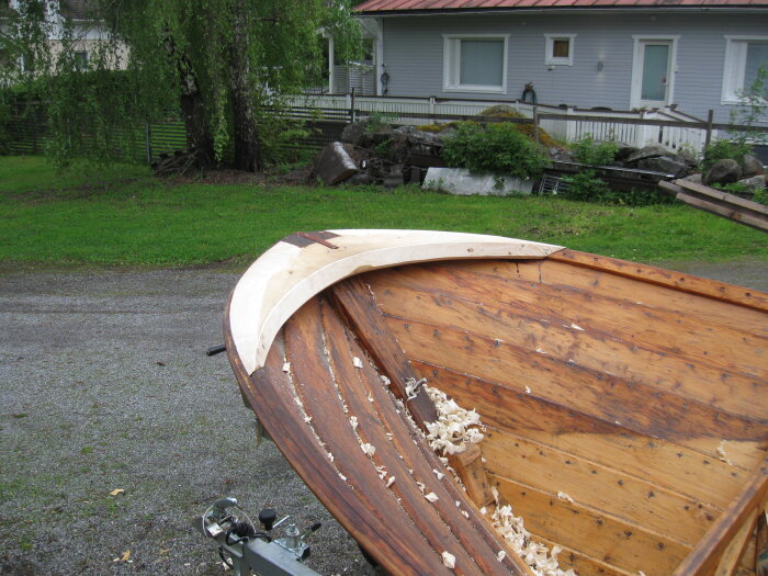 En halvfärdig träbåt under renovering med träspån kvar i båten, bredvid ett hus.