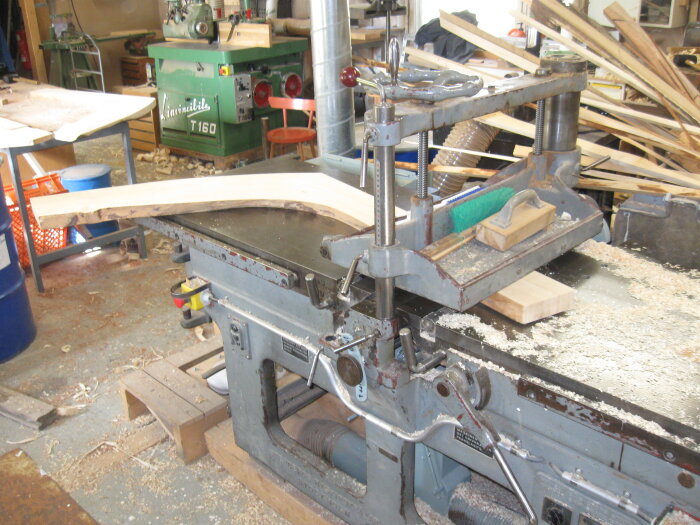 Verkstadsmiljö med stor hyvelmaskin och träbitar, omgiven av snickarbänkar och verktyg. Träspån strödda runt maskinen.