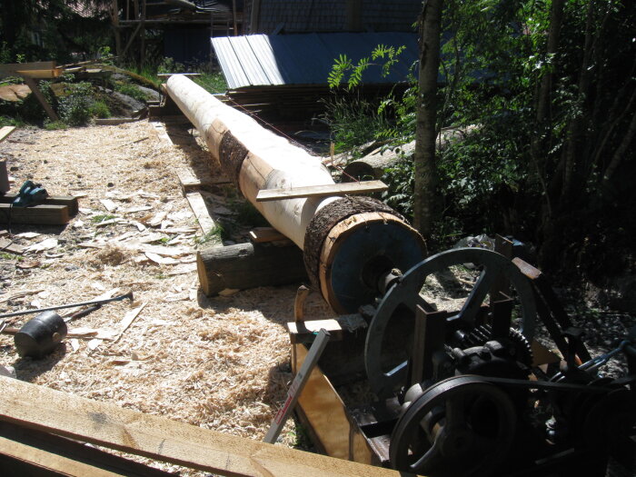 Stort trästycke sätts upp för bearbetning med hjälp av en hemmabyggd maskin i ett utomhusarbete, omgiven av träflisor och gröna träd.