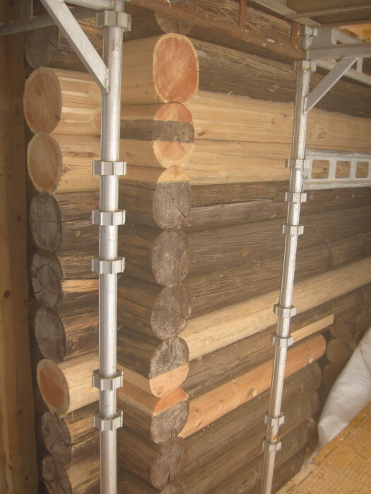 Timmerstomme under reparation med ställning, synliga stockar och metallstöd. Nytt och gammalt timmer syns i väggen.