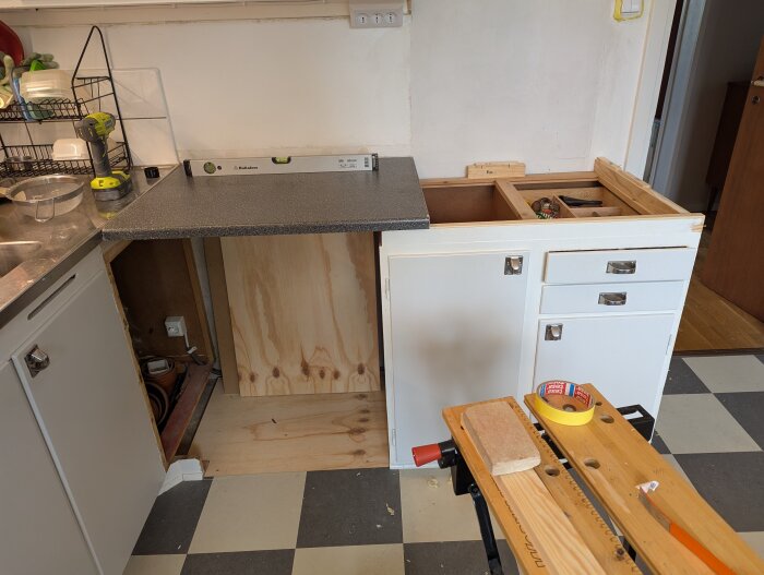 Kök med vitrin- och underskåp, pågående bänkbygge med ny bänkskiva, nivåpass och verktyg synliga på skivor.