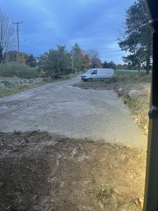 Grusyta bredvid ett hus vid en väg, med en parkerad vit skåpbil och omgivande natur.