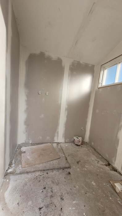 Renovering av badrum med betonggolv och väggar i gips. Ett skydd täcker golvbrunnen. Fönster och naturligt ljus syns i bakgrunden.