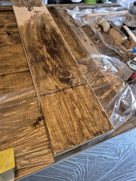 Bräda med ojämn brun betsning, delvis missfärgad, liggande på plasttäckta ytor med verktyg och burkar i bakgrunden.