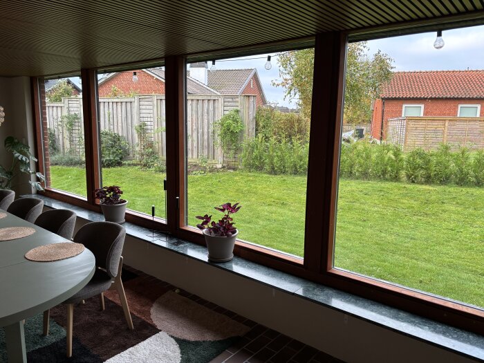 Rymligt rum med stora fönster, som släpper in mycket ljus, utsikt mot en grön trädgård och modern inredning med fåtöljer och en matta.