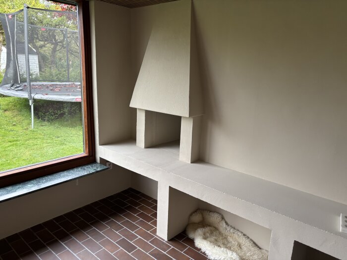 Braskamin med murad inramning i ett rum med fönster mot trädgård och studsmatta. Bruna klinkergolv och fäll på golvet framför kaminen.