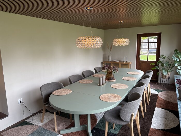 Modernt matrum med grönt ovalt matbord, åtta gråa stolar, två hängande lampor och matta. Växter och dekorationer kompletterar rummet.