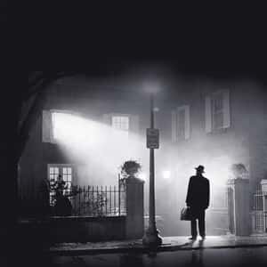 En man med hatt står framför ett hus upplyst i natten, med en väska i handen, under en gatulampa.