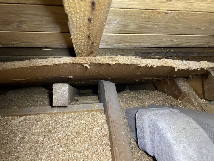 Vindsutrymme med träbalkar och ett fack delvis fylld med sågspån, visar en oisolerad sektion jämfört med intilliggande fylldt område.