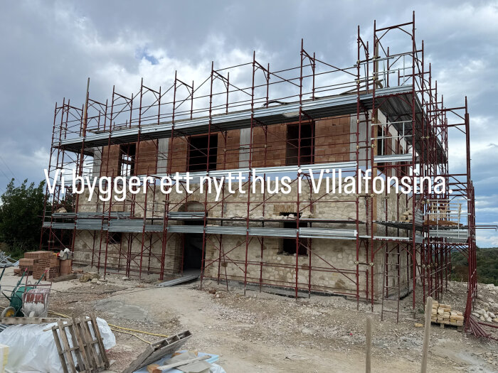 Byggarbetsplats med stenhus under uppförande omgärdat av byggnadsställningar, texten "Vi bygger ett nytt hus i Villalfonsina" syns på bilden.