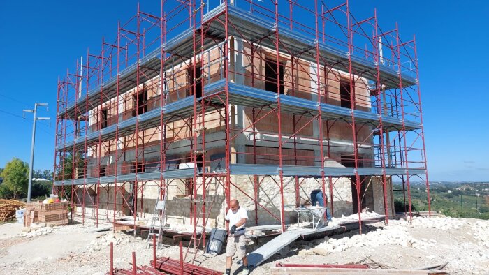 Byggarbetsplats med en flerplansbyggnad omgiven av byggnadsställningar. Person i förgrunden som arbetar, medan en annan person är vid ställningen.