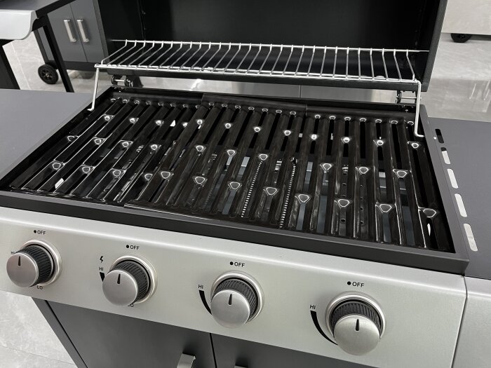 Gasolgrill med öppet lock och kontroller för värmeinställning. Grillgaller i blankt material, eventuellt gjutjärn eller plåt.