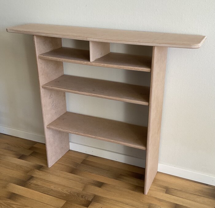 En odefinierbar möbel med hyllor av mdf, delvis färdigställd, bred toppskiva med kurva och ekkantlist, stående mot en vägg i ett rum.