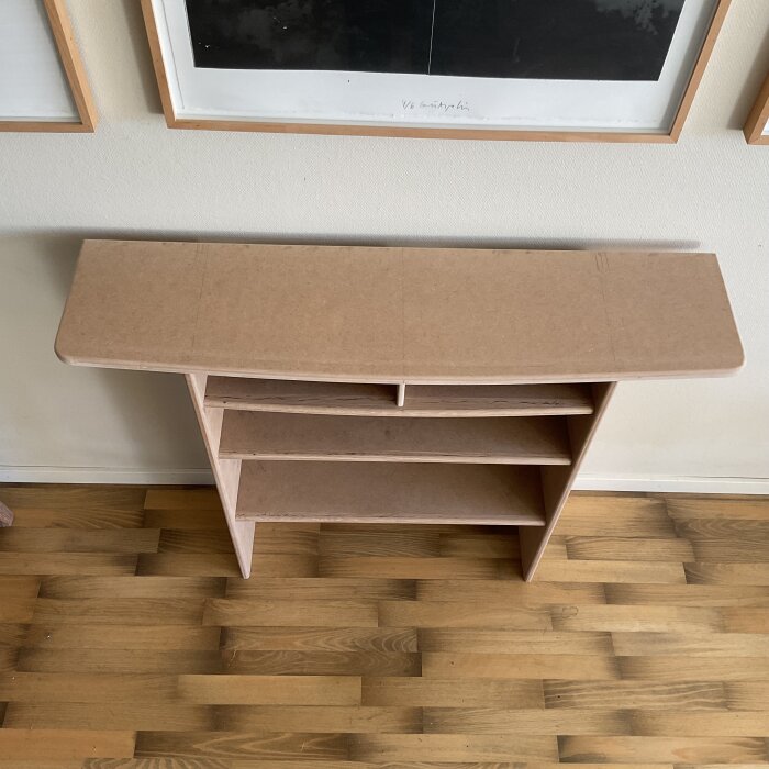 Omonterad möbel av MDF med toppskiva och hyllor med svag kurva, placerad mot en vägg framför tavlor på ett trägolv.