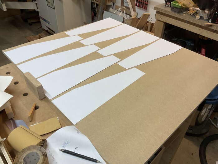 Pappersmallar av olika former utlagda på ett arbetsbord i verkstad, omgivna av verktyg och material, för förberedelse av mdf-bitar.