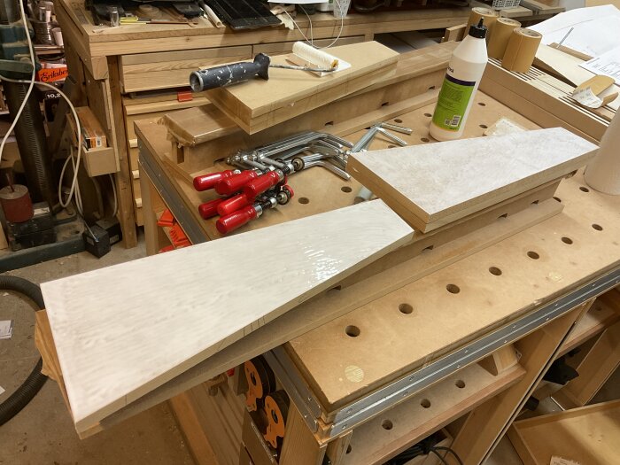 En arbetsbänk i träverkstad visar limmade MDF-bitar med röda tvingar och limflaska. Verktyg och material syns i bakgrunden.