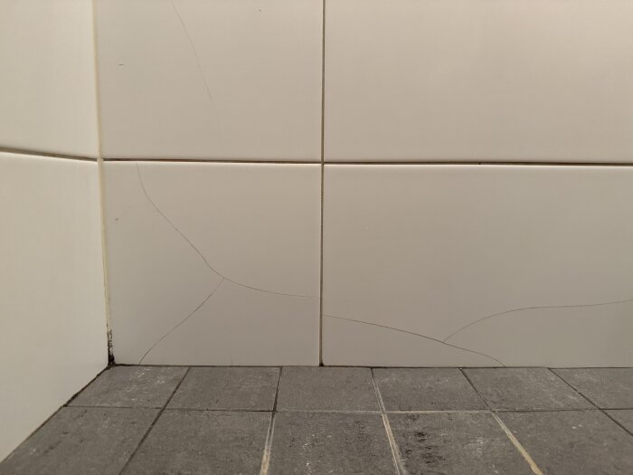 Spickor i duschplattor vid golvets hörn, med synlig mörk missfärgning i nederkanten. Plattorna är vita och golvet mörkt.