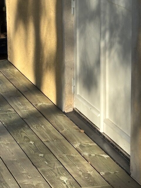 Terrassgolv i trä med distanser vid putsad fasad, nära en vit dörr. Sol-mönstrade skuggor syns på fasaden. Gångjärn på dörren synliga.