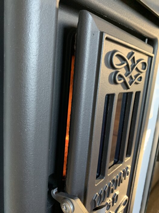 Närbild av en svart vedeldad kamin med dekorativa detaljer på dörren, där ett orange sken av eld syns inuti.