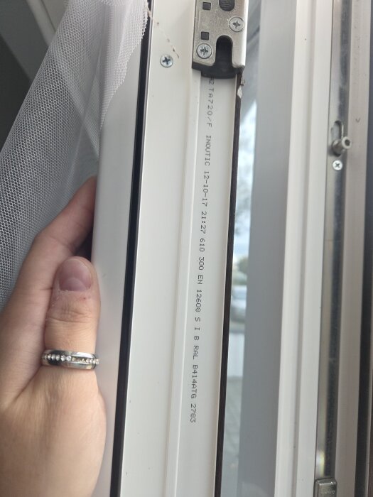 Närbild av PVC-fönsterkarm med fokus på övre gångjärnet och en hand som håller fönstret, synlig text och skruvar.
