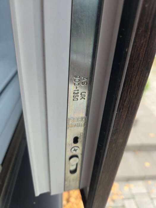 Metallbeslag på PVC-fönstrets gångjärnssida, med text "AS UK 900-1350" och olika justeringsmarkeringar synliga.