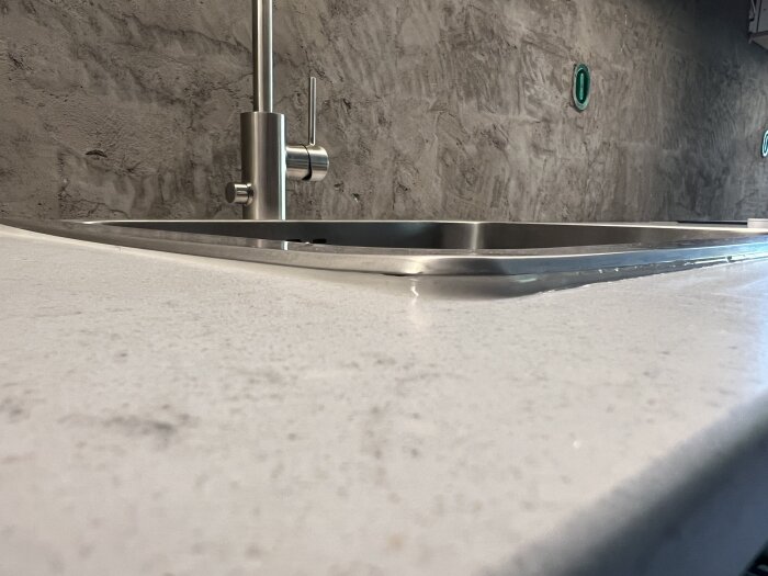 Installerad diskho inte tillräckligt nedtryckt i bänkskivan, med synlig kant. Syns även en modern kran och grov textur på väggen i bakgrunden.