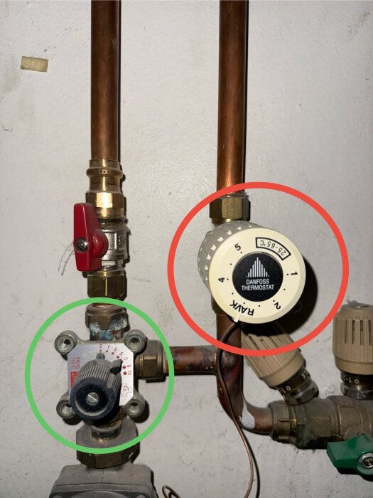 Termostat med två justerbara reglage markerade med röd och grön cirkel på kopparrör i ett VVS-system.