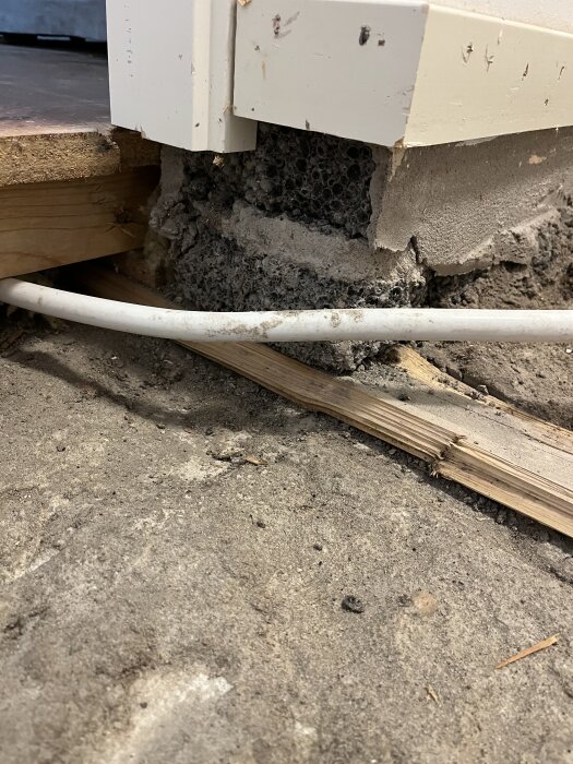Ojämnt betonggolv med träregel under en källarvägg, visar rör och ojämnheter vid golvkant nära väggen.
