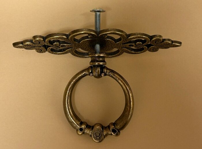 Antikt mässingsbeslag med dekorativ platta och dragring, fäst med genomgående skruv, för en byrålåda.