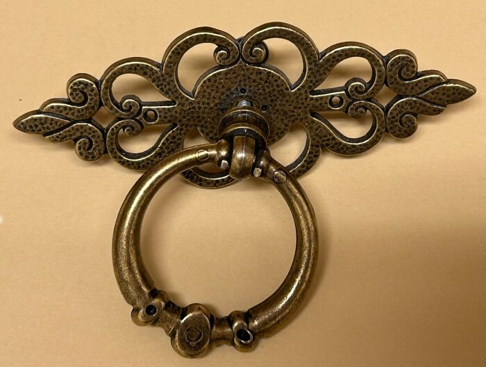 Antikt bronserat lådbeslag med ringhandtag och detaljerad bladdekor, monterat med en genomgående skruv och mässingspik.