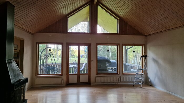Vardagsrum med furupanelat tak, stora fönster och stege. Väggarna har tygtapeter. Armaturer har tagits bort. Utsikt mot trädgård.