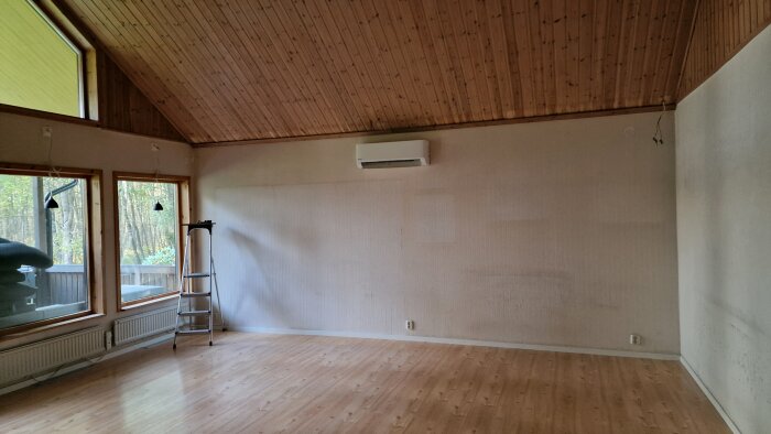 Vardagsrum med träpaneltak, stora fönster och beige väggar utan dekoration. En stege vid väggen, luftkonditionering ovanför.