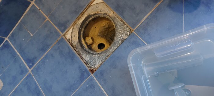 En sned golvbrunn med lös täckram och fuktskadade fogar i en tvättstuga med blå kakelplattor.