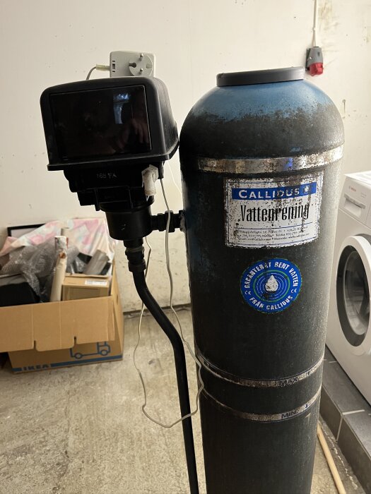 Callidus vattenfilter från 1998 med behållare för kaliumpermanganat, stående i ett garage, intill en kartong och en tvättmaskin.