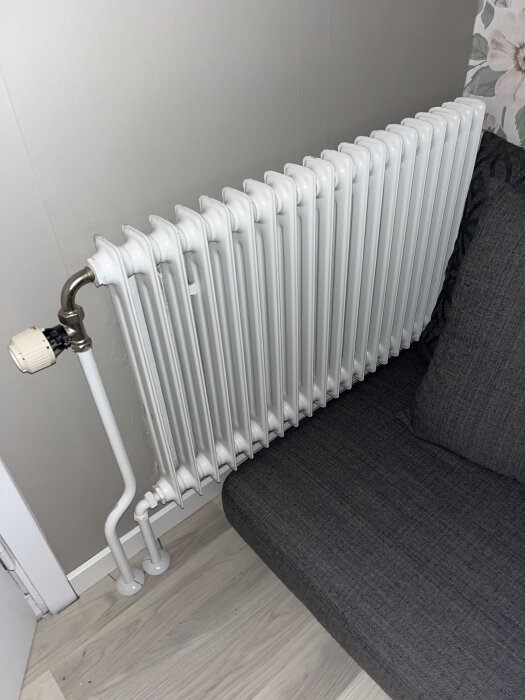 Vit sektionsradiator på ljus vägg bredvid grå soffa, med termostatventil och röranslutning till golvet.