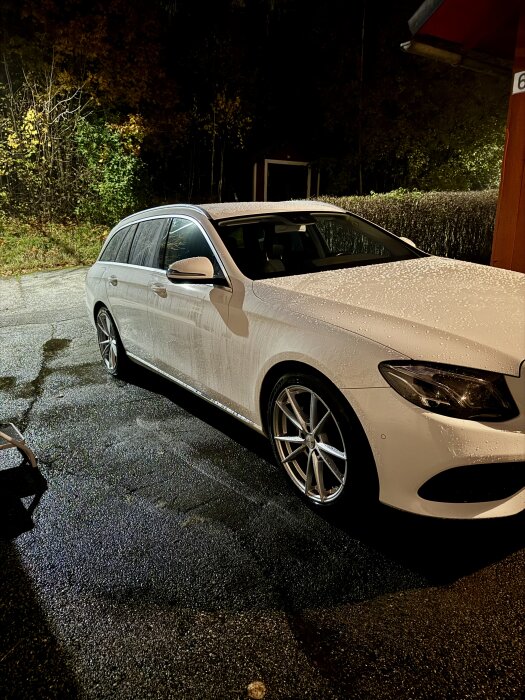 Vit bil med vattenpärlor efter tvätt, står parkerad på en våt asfaltsyta omgiven av träd.